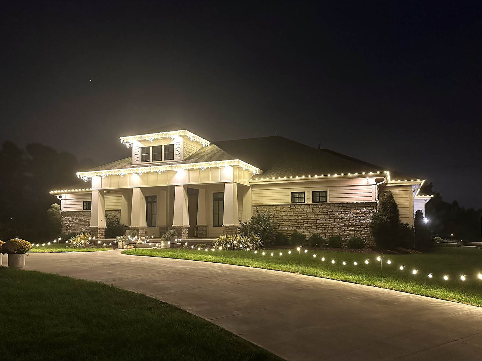 Estate home with dormer and columned entry, roofline lit and driveway lined with bistro lights